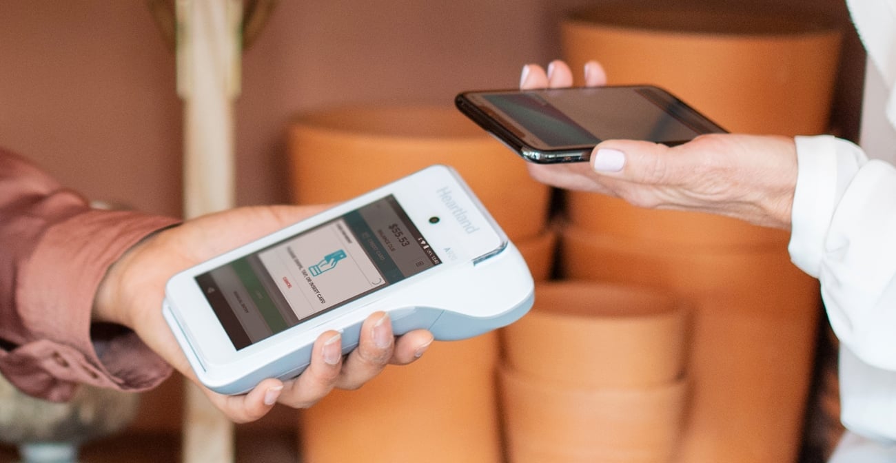 Close-up of two hands: one on left side holds mobile point of sale device, one on right side holds smartphone