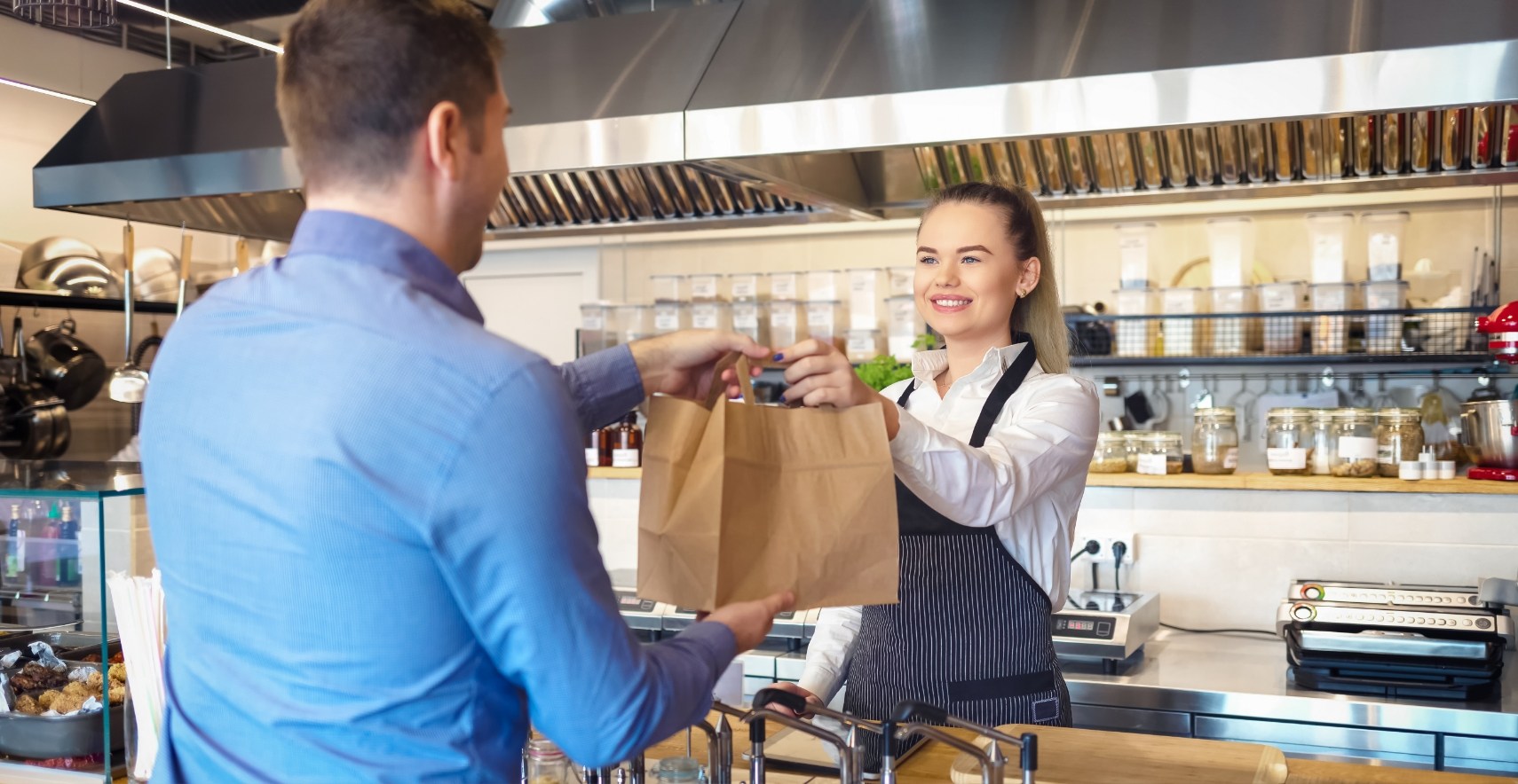 Employee in apron handing takeout paper bag to customer across counter inside restaurant