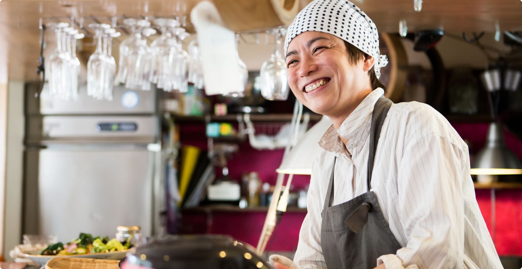 Person wearing chef apron and patterned bandana smiling broadly in professional kitchen setting