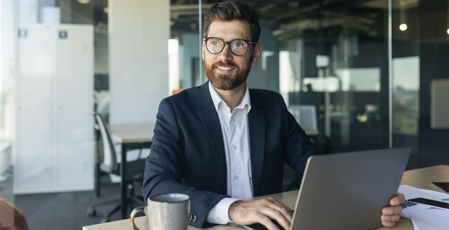 A man sits in a glass office, working on his laptop with a coffee mug in front of him.