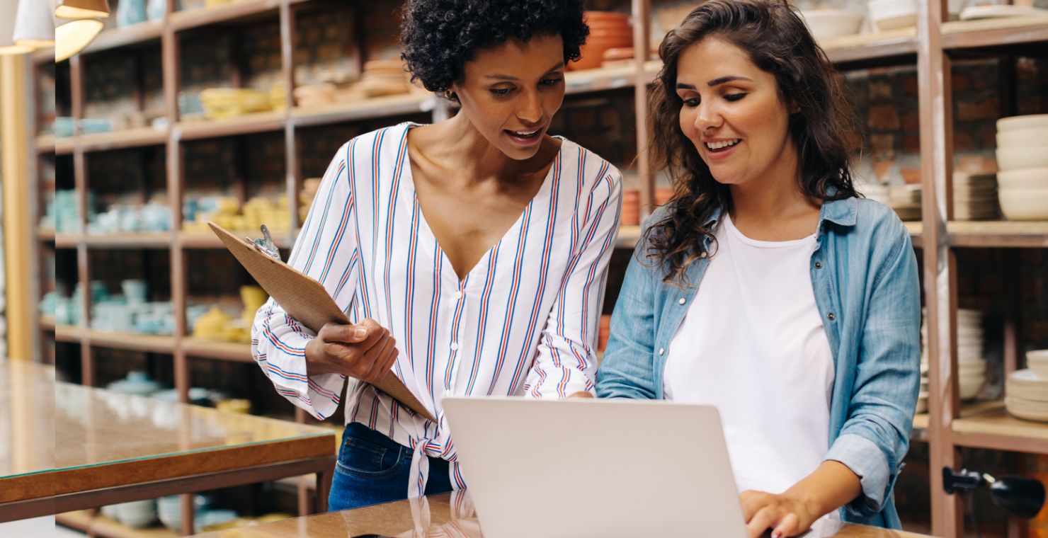 Two women from a small business analyze data on a laptop with inventory shelves behind in the background.