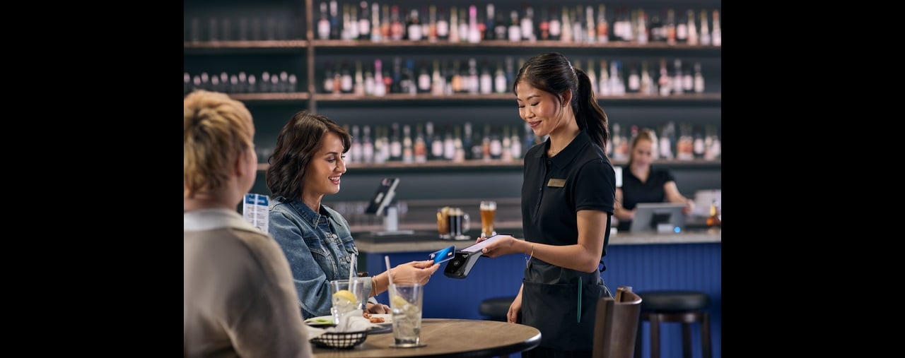 Smiling server holds payment device as seated customer taps credit card to pay in bar-and-grill setting