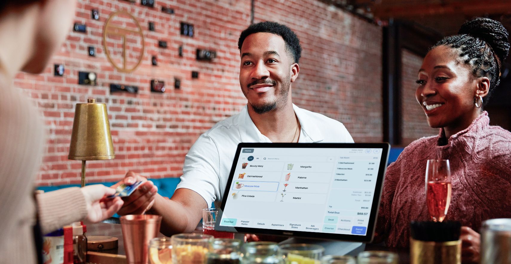 Two people smile as one of them hands payment card to restaurant employee who stands near point of sale system