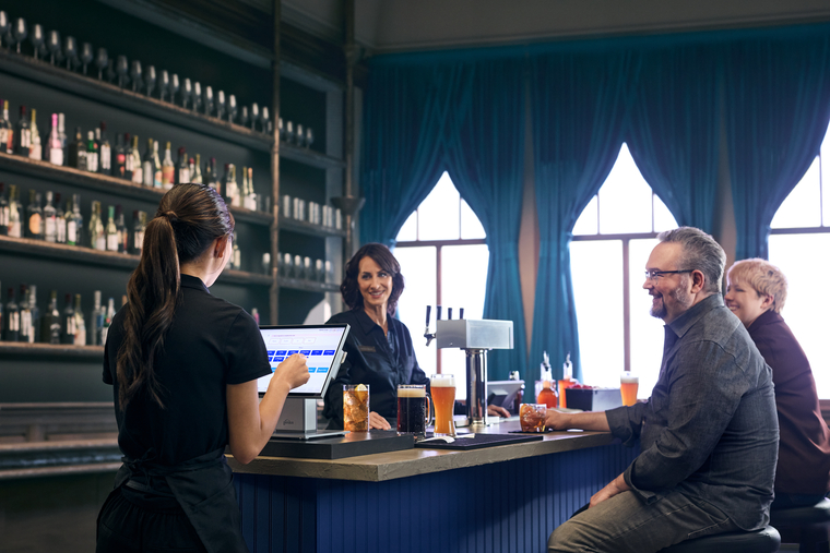 Server using countertop device to enter order in bar area of restaurant near bartender and customers