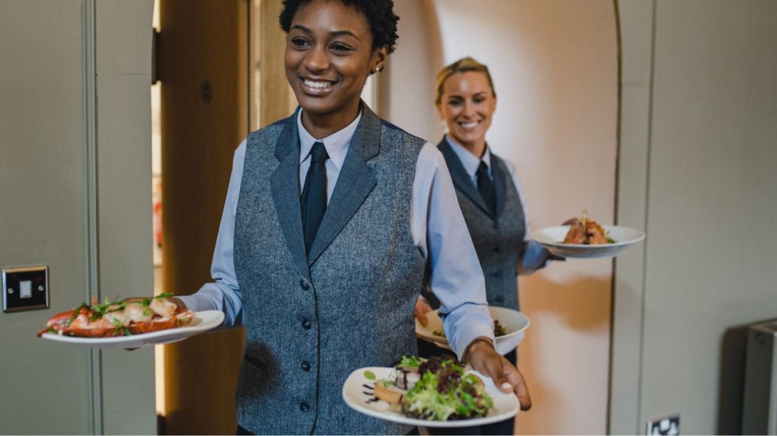 Two smiling servers walking out of a restaurant kitchen holding plates of food