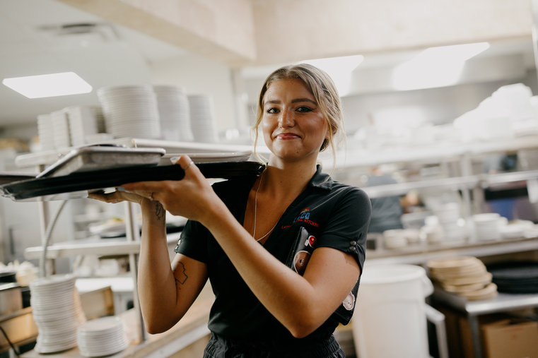 Smiling restaurant worker in dark uniform balancing service tray inside commercial kitchen