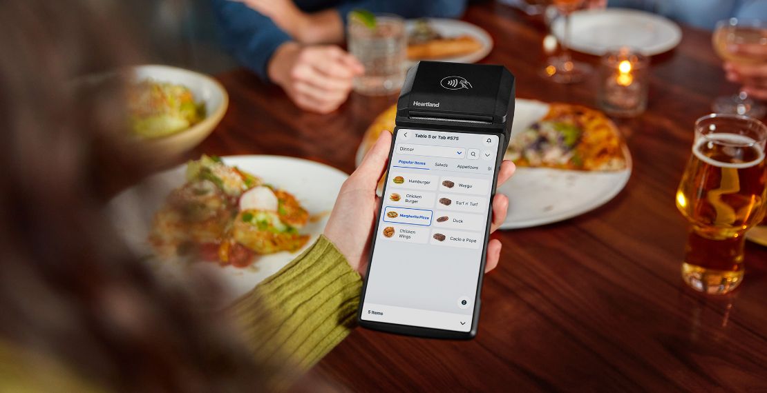 Overhead shot of someone holding Heartland Mobile POS with tableside ordering screen over a restaurant table of food
