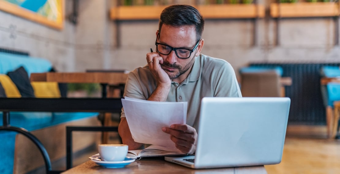Man in coffee shop sits in front of laptop and studies information on a stack of papers.