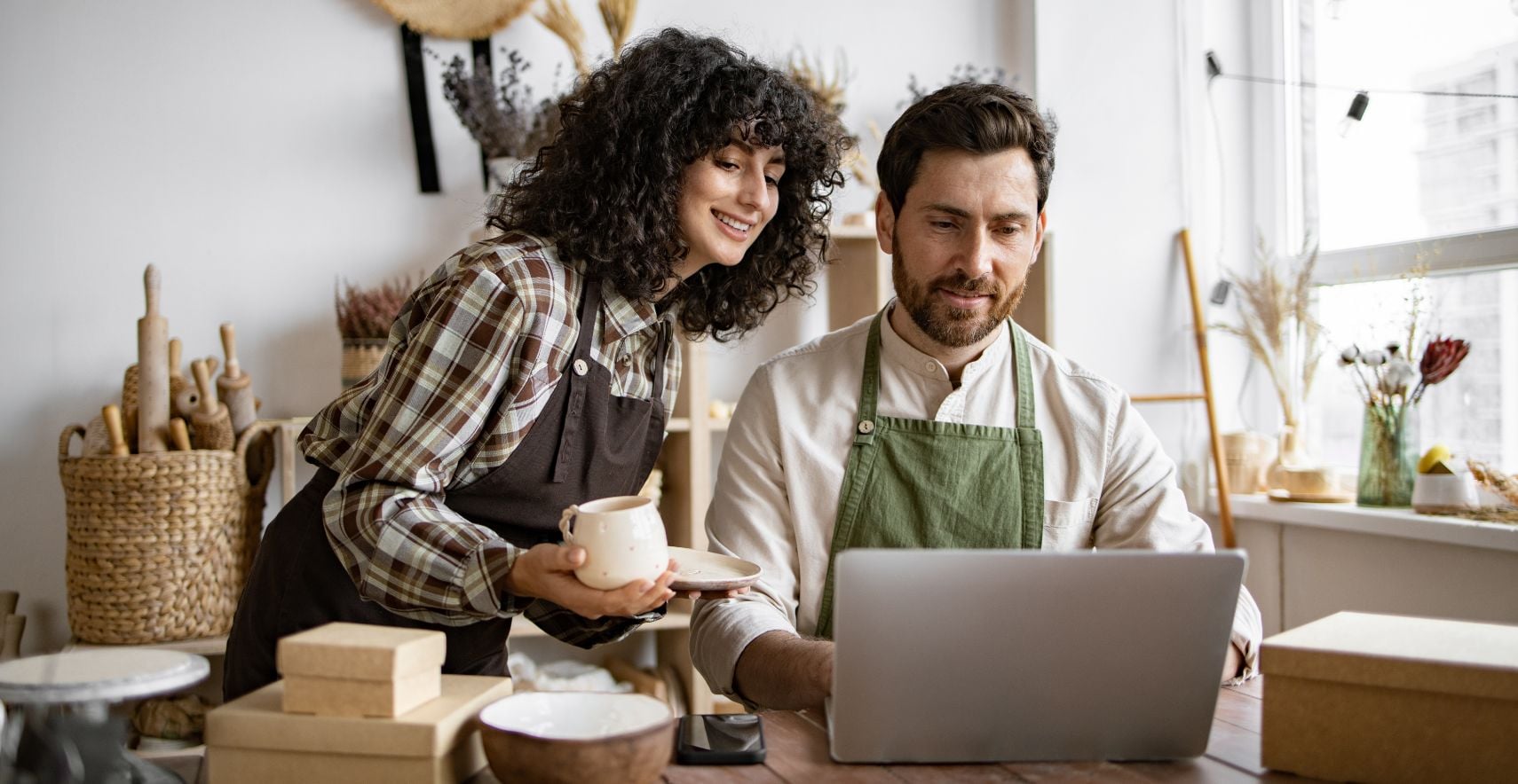 Seated retailer works on laptop as standing coworker looks on while holding coffee mug and saucer