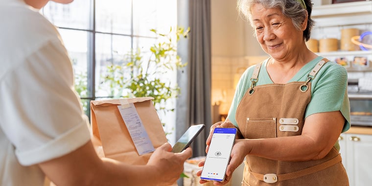 A small business owner hands a smartphone to a customer at a counter, demonstrating a mobile payment app.