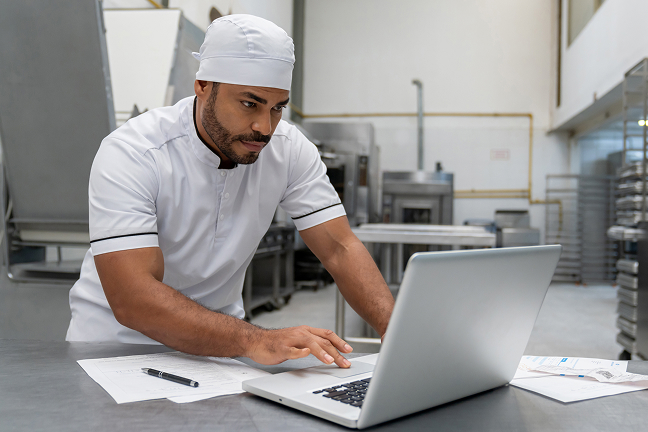 Person in chef's uniform using laptop in restaurant kitchen