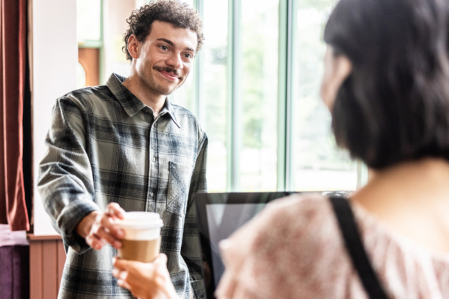 Smiling person handing disposable coffee cup to customer across counter