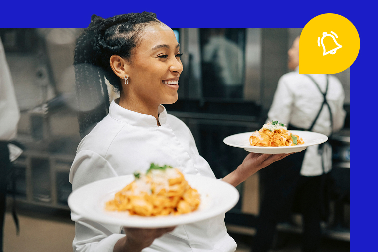 Smiling person in chef's uniform holding two plates of prepared food in commercial kitchen