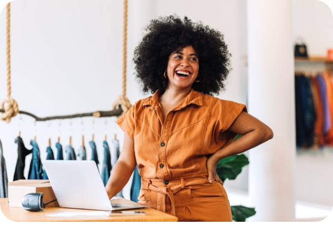 Smiling retail business owner standing behind a checkout counter with a laptop and clothing racks in the background.