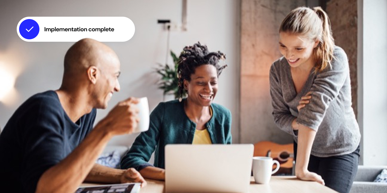 Image of three smiling people sharing a laptop with ‘Implementation complete’ text overlay