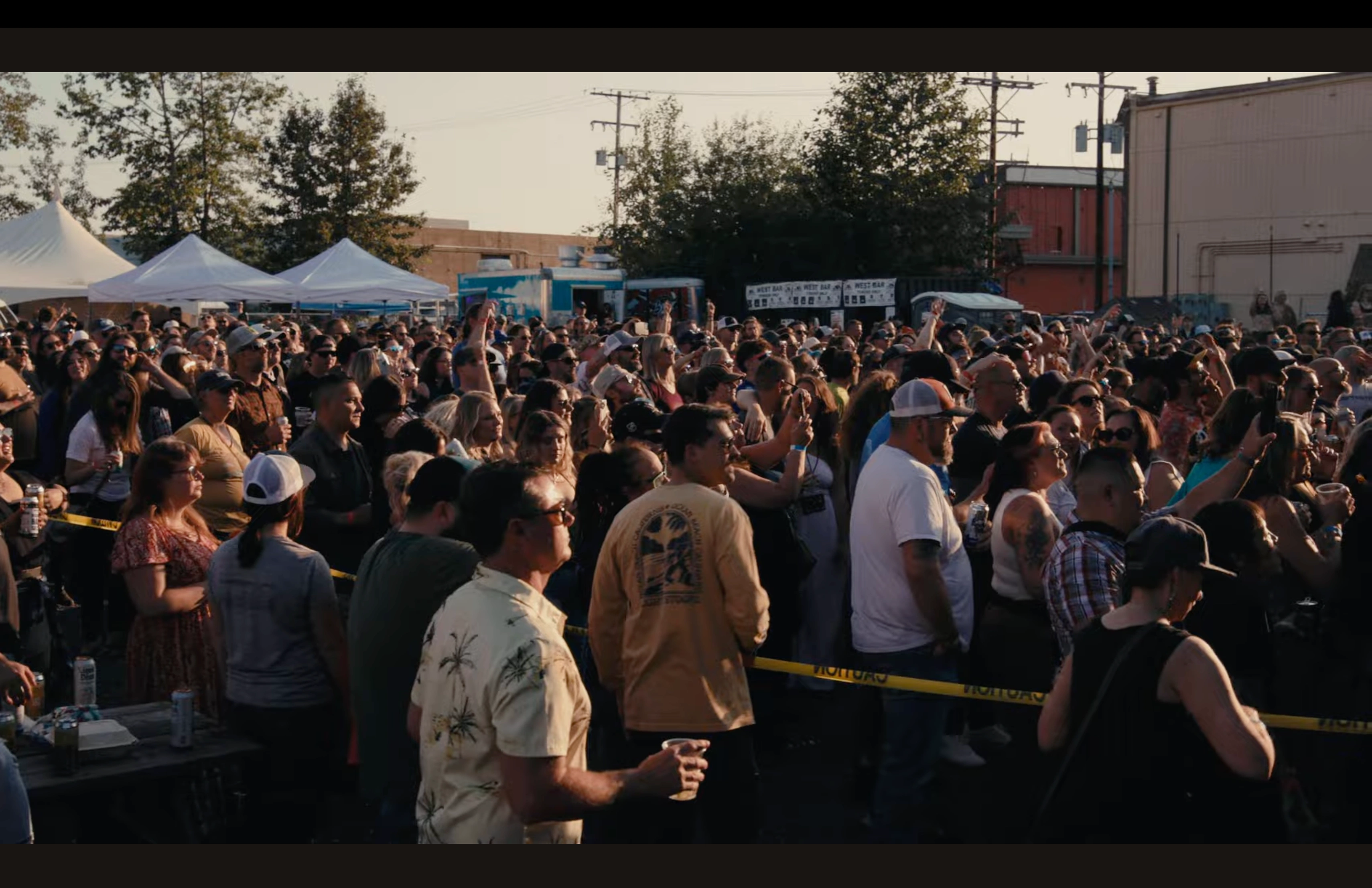 A crowd of people stand in an outdoor concert venue at sunset, listening to music.