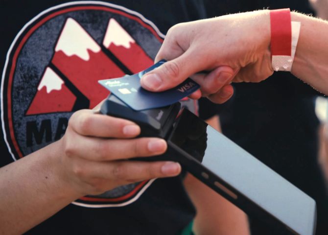 A closeup of a hand holding a mobile payments device and another hand tapping a payment card on the top of the mobile device.