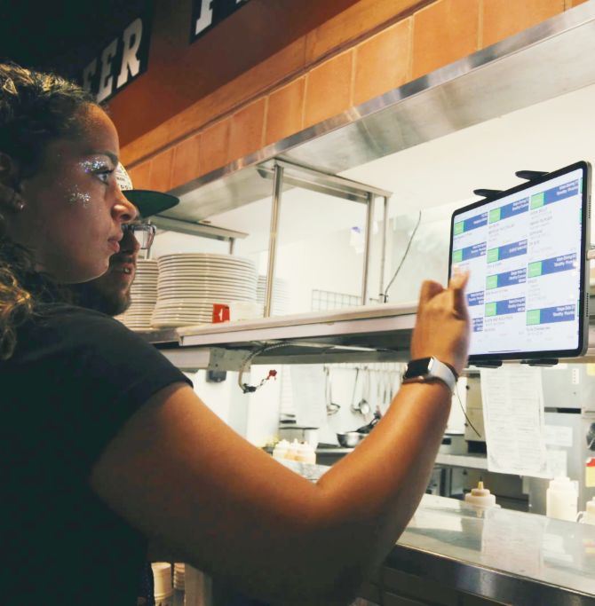 A woman in the back of house at a restaurant taps a screen attached to a shelf.