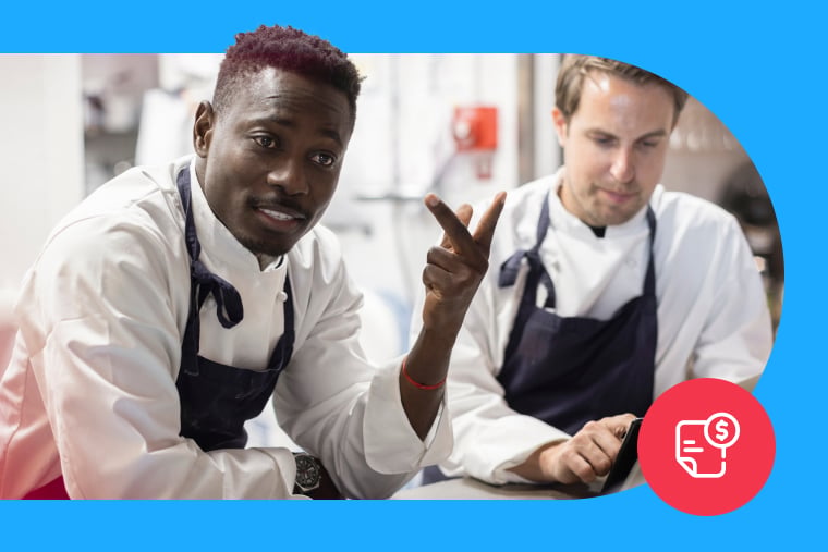 Two restaurant staff wearing black aprons work in a commercial kitchen.