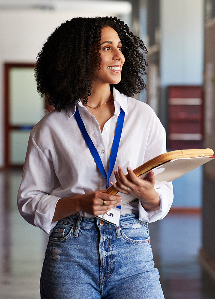 A higher education staff member holds a laptop and folder while walking down a school hallway smiling.
