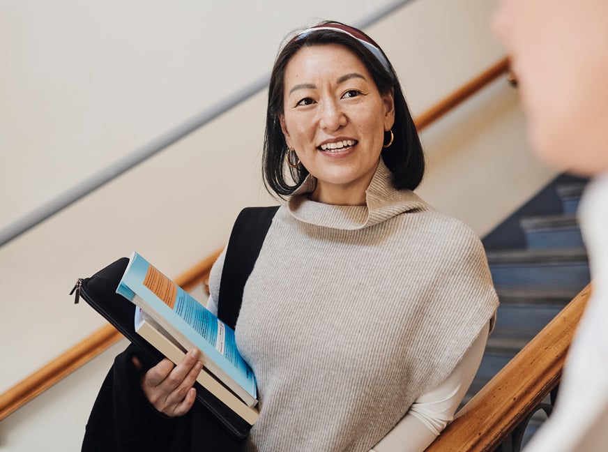 Person in a higher education setting holds books in a hallway while speaking with a fellow student.