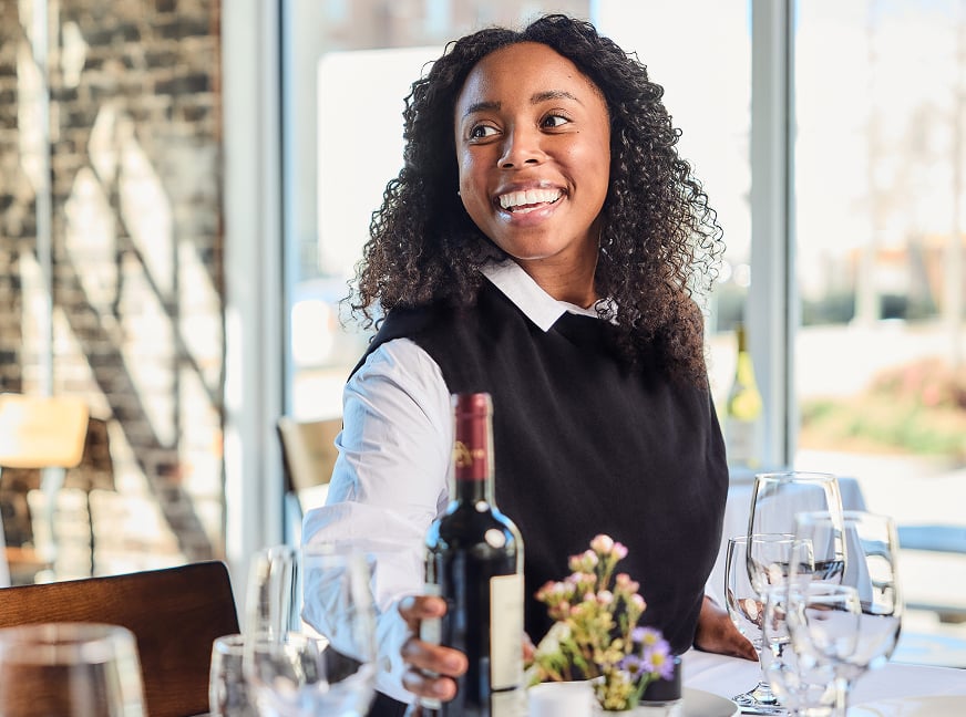 Smiling hospitality worker places wine bottle on a table set with wine glasses.