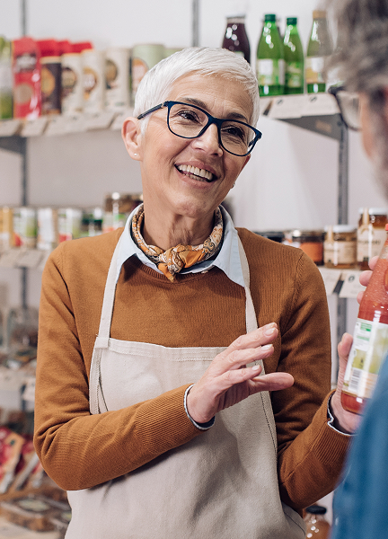 Retail worker stands in front of shelves stocked with food and drink products while telling a customer about one of them.