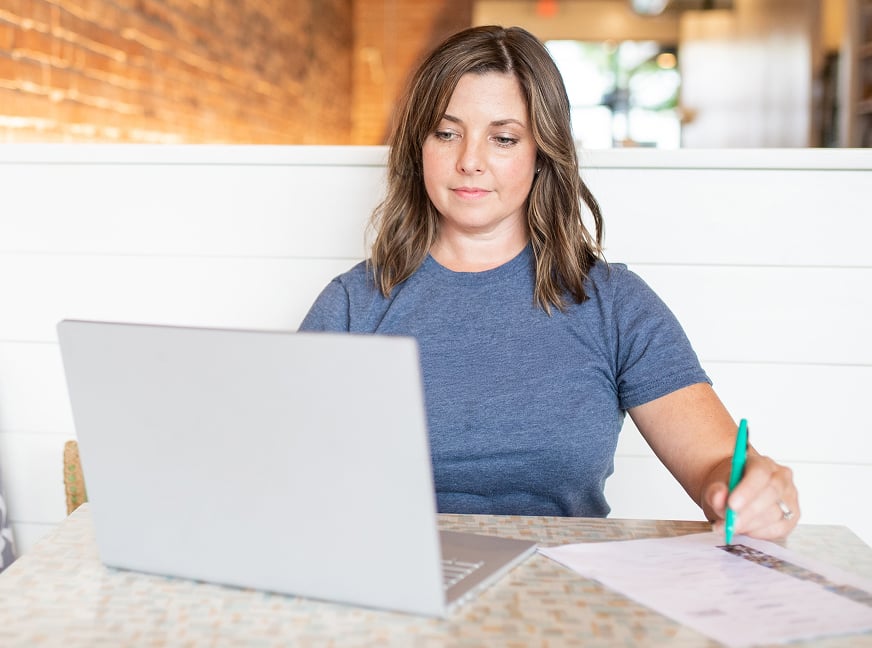 Small business owner sitting at a table makes notes on paper while looking at their laptop.