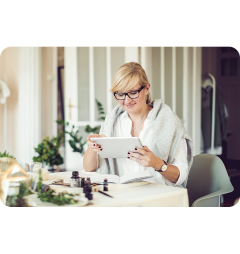 Retail worker wearing glasses and white top holds tablet while seated at table with small bottles