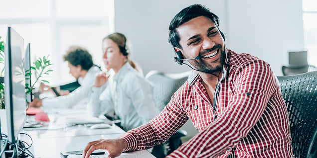 Man wearing a headset and red checkered shirt laughing at his computer in a call center.