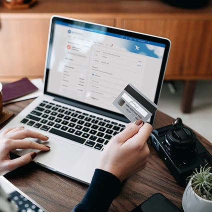 A person holds a credit card while booking a flight on a laptop at a wooden desk.