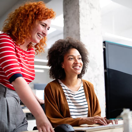 Two smiling female colleagues working together at a computer monitor in an office.