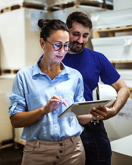 Woman using a tablet alongside a worker holding a hard hat in a warehouse.