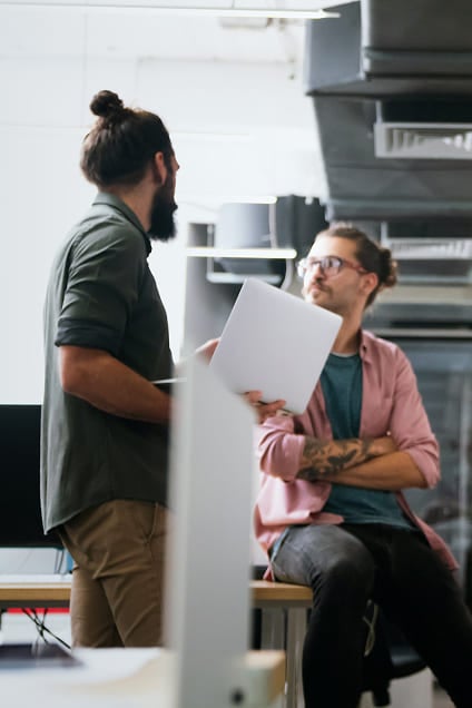 Man standing with a laptop talking to a colleague sitting on a desk.