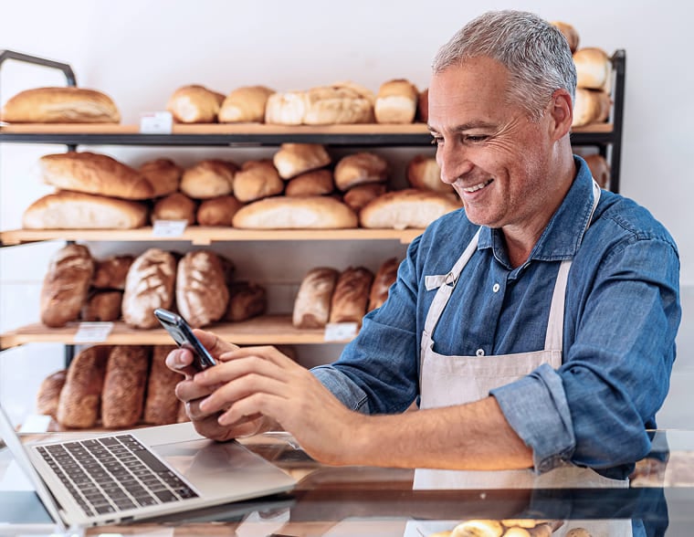 Middle-aged man in an apron using a smartphone in a bakery with bread shelves behind