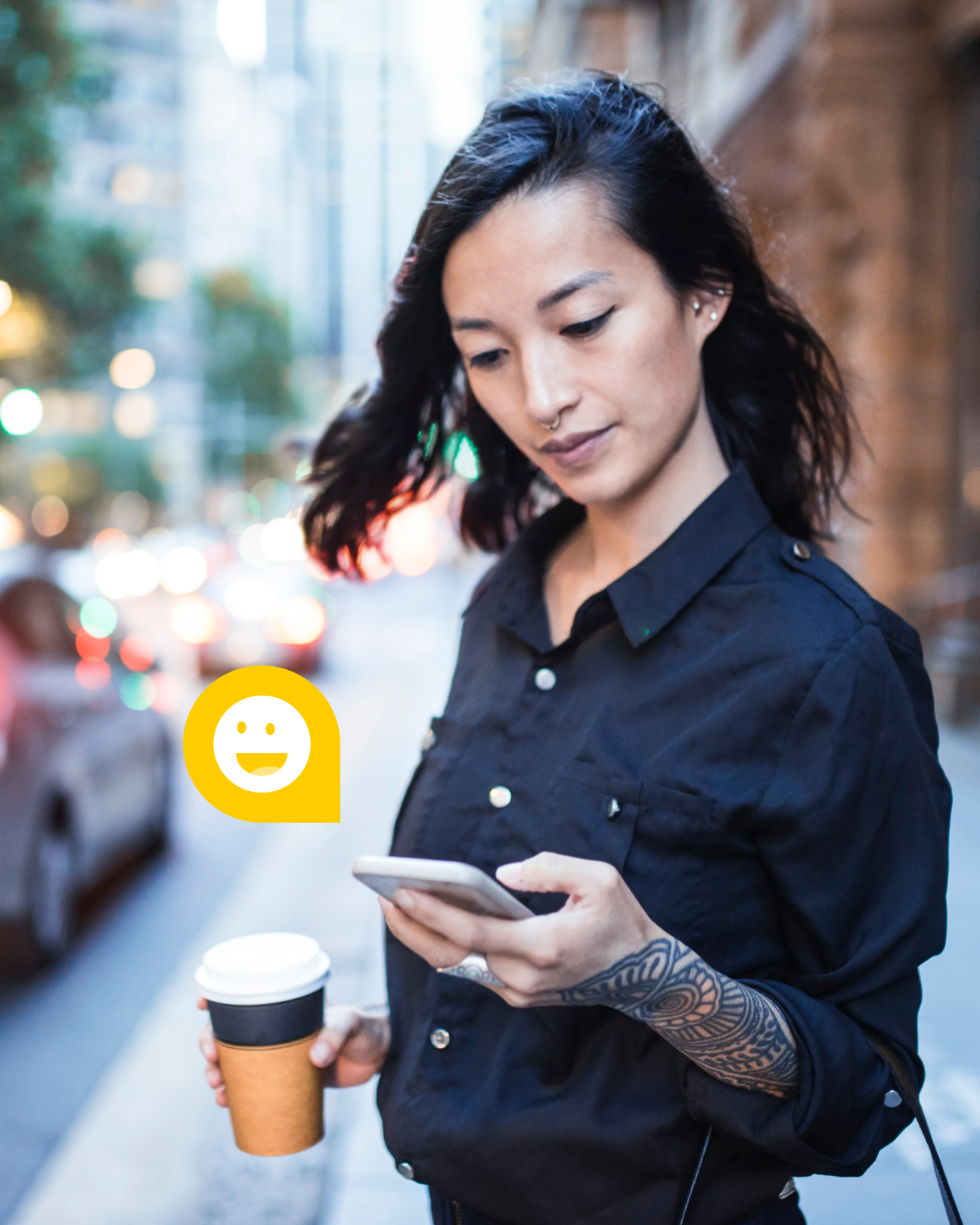 Woman on street looking at mobile device while holding coffee. Smiley face emoji is on the screen.