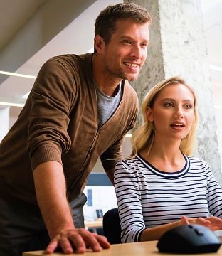 A man leans over a woman sitting at a desktop computer.