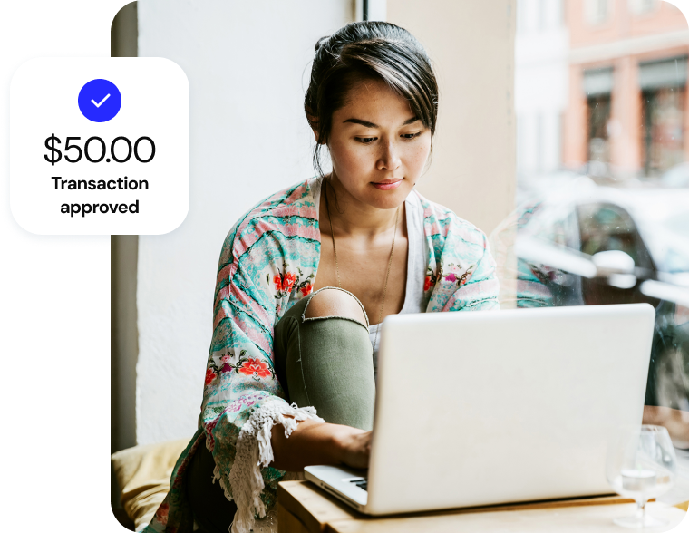 A woman works at her laptop in a bright coffee shop, and a soft UI callout sits in the foreground reading “$50.00, transaction approved.”