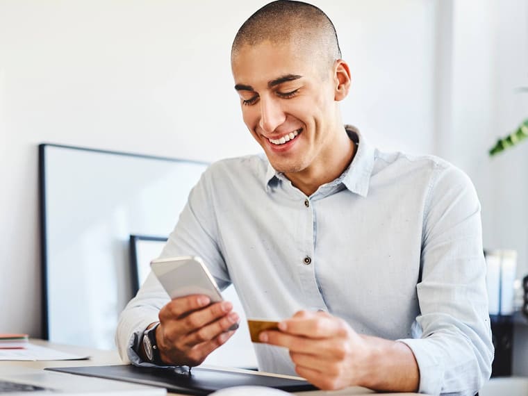 Man holding a credit card and smartphone while sitting at an office desk.