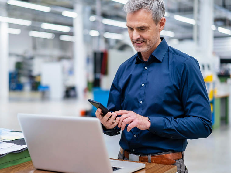 Man in a blue shirt checking his phone at a laptop workstation in a warehouse.