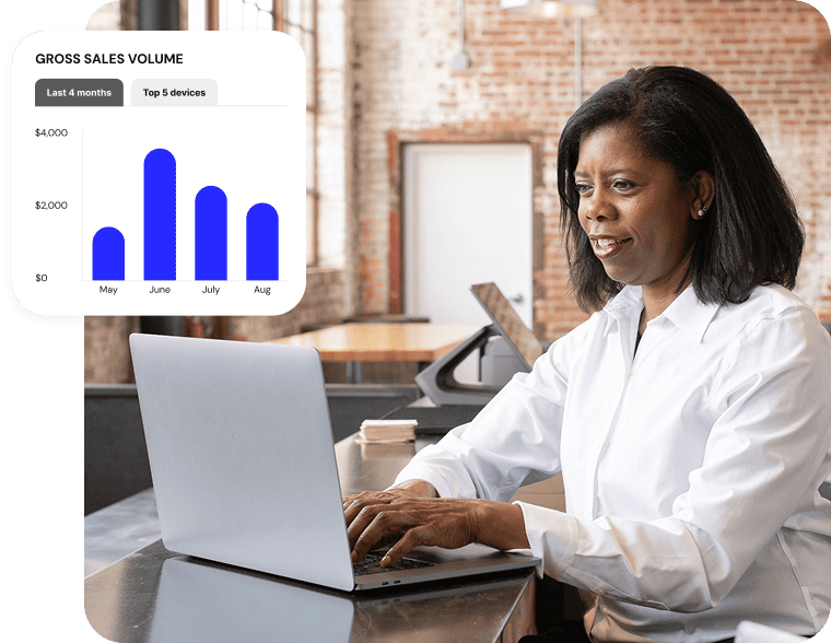 Female employee sitting at a desk typing on an open laptop. A popup UI screen shows a gross sales volume report from the back office software.