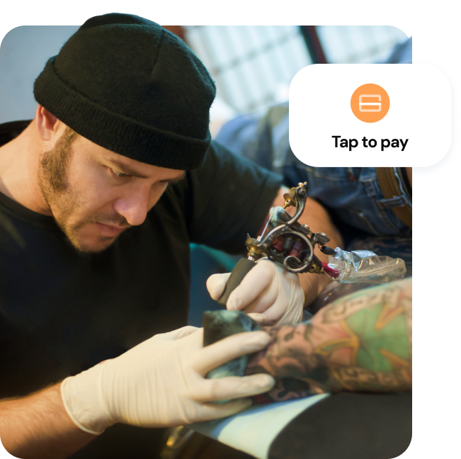 A tattoo artist applying ink to a customer’s forearm with a “Tap to pay” overlay indicating payment capability.