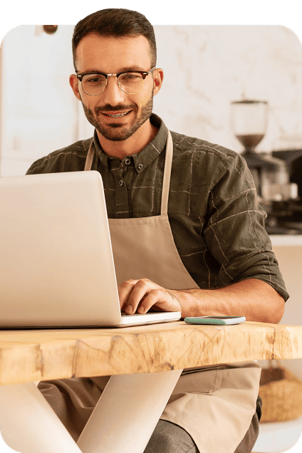 Un homme à lunettes et à barbe, portant un tablier, travaille sur son ordinateur portable.