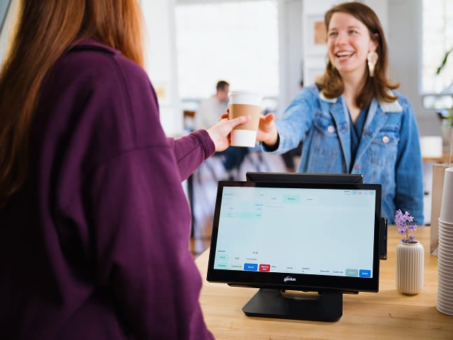 Femme recevant un gobelet de café à emporter au-dessus d'un comptoir de caisse équipé d'un terminal de point de vente à écran tactile.