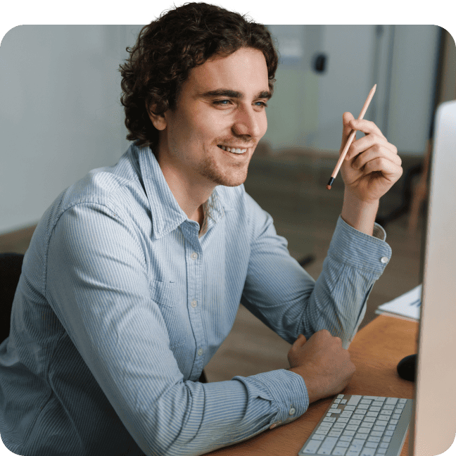 Man in a light blue shirt holding a pencil and looking at a computer monitor.
