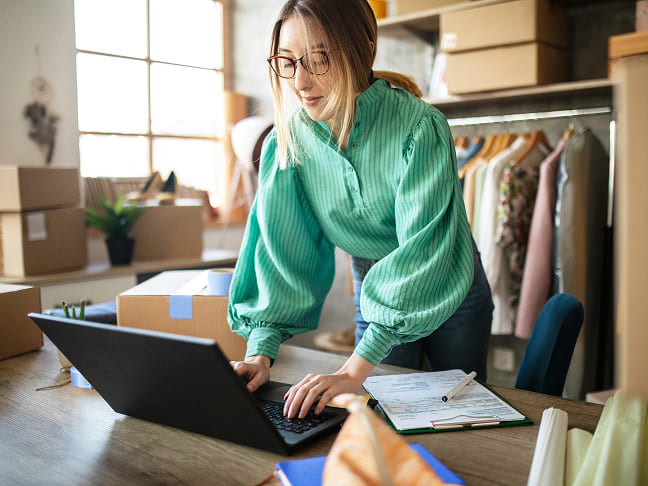 Woman wearing glasses and a green top typing on a laptop surrounded by shipping boxes.