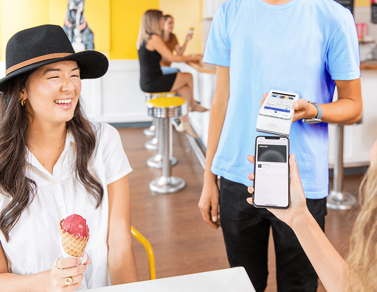 A customer at an ice cream shop eats an ice cream cone while her friend pays for it on a card machine from a phone mobile.