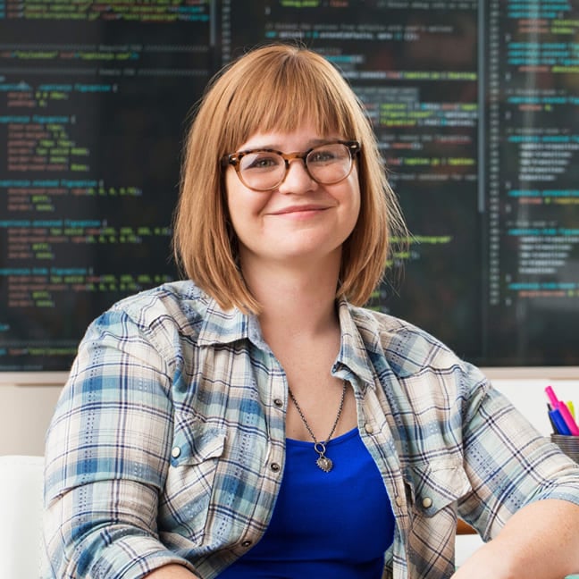 A woman with glasses and a plaid shirt smiles while sitting in front of a screen displaying lines of computer code.
