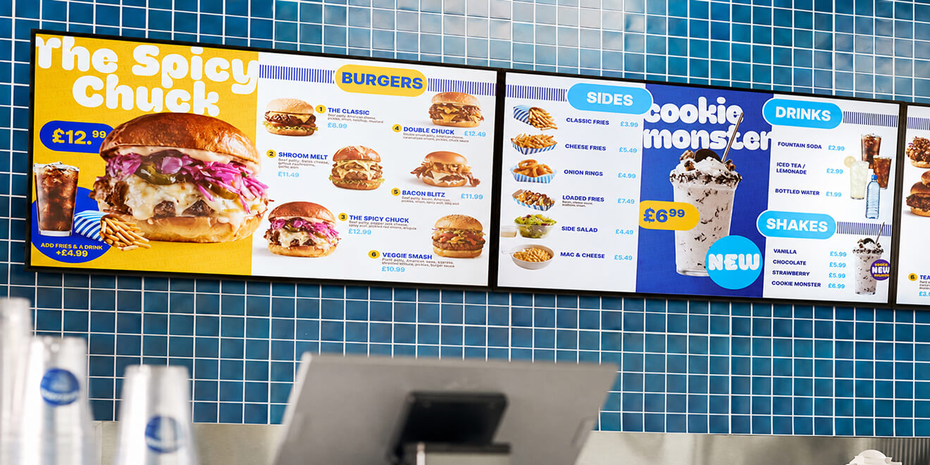 Digital menu boards in a fast-food restaurant showing burgers, sides, drinks, and shakes, with a large “Spicy Chuck” burger featured on a blue tiled wall.