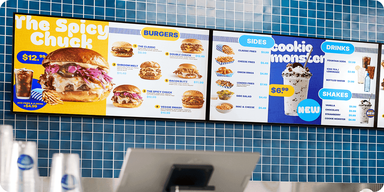 Digital menu boards in a fast-food restaurant showing burgers, sides, drinks, and shakes, with a large “Spicy Chuck” burger featured on a blue tiled wall.